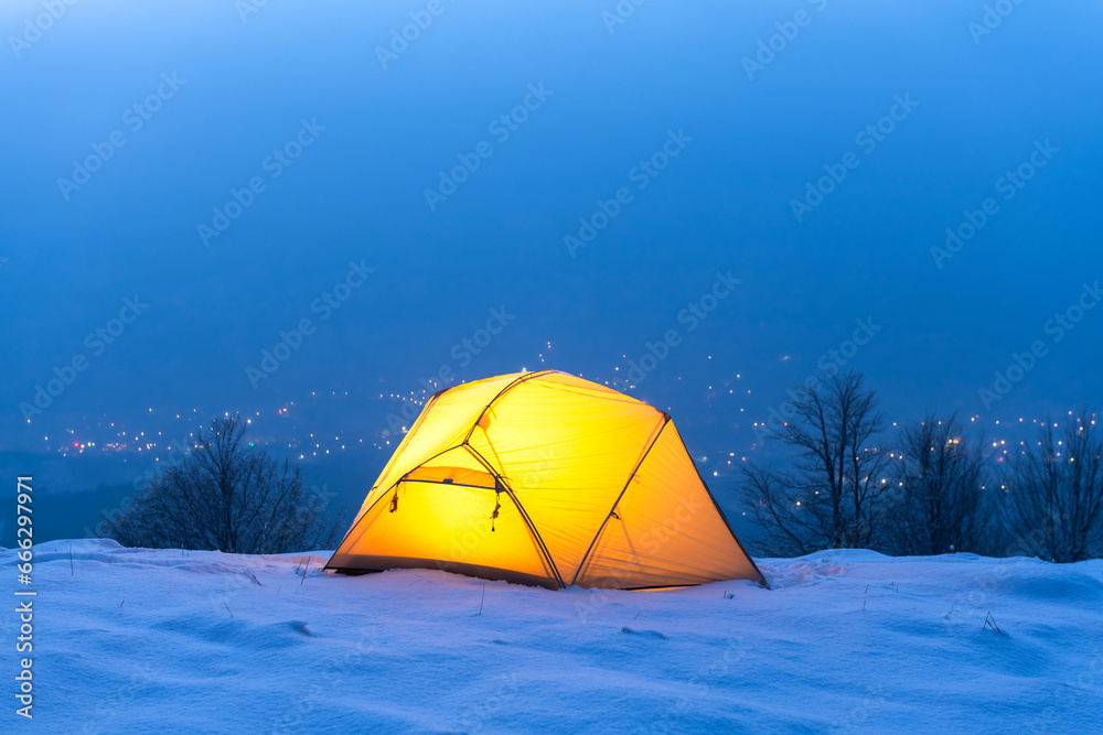 A yellow tent illuminated from within, set amidst the enchanting glow of city lights. A portrayal of travelers camping in the winter mountains, embodying the essence of travel and exploration