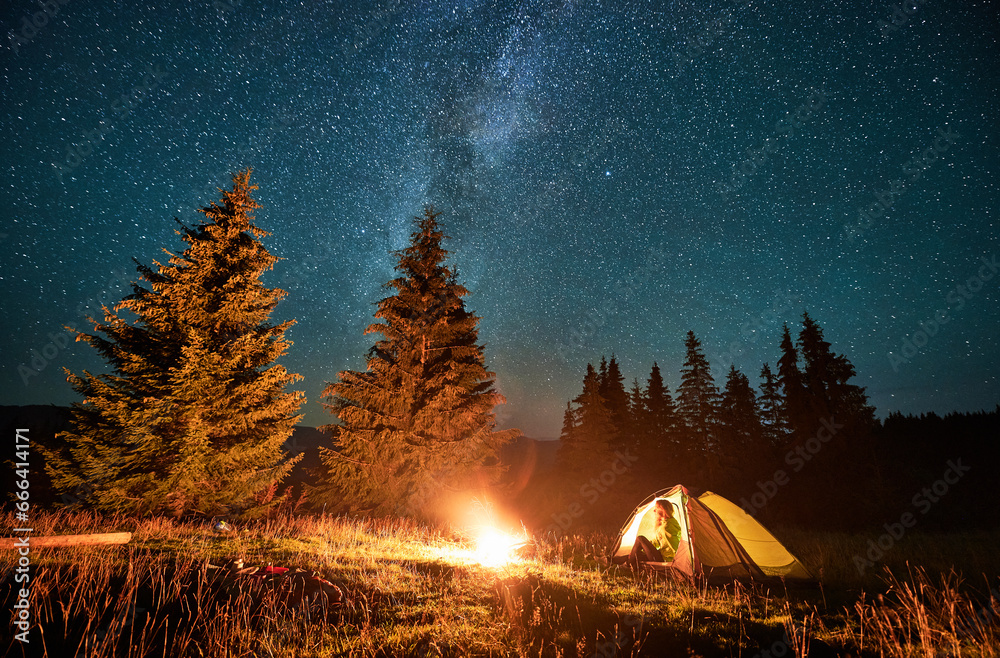 Night camping in mountains under starry sky and Milky way. Female tourist sitting in tent in campsite, admiring landscape and nature, fire burning. Concept of traveling and hiking.
