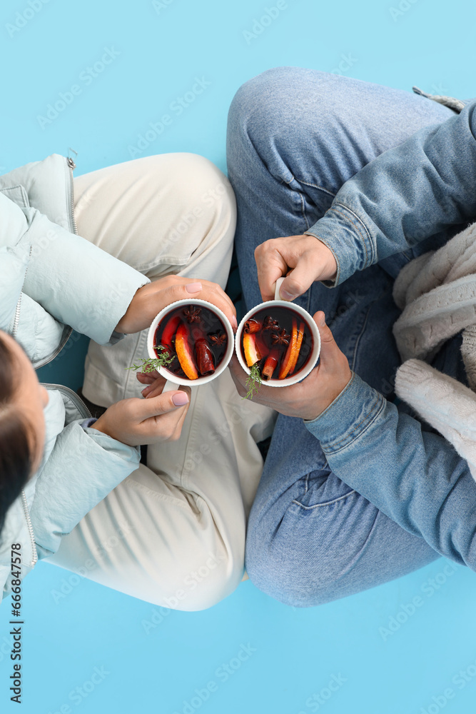 Couple in warm clothes with mugs of tasty mulled wine sitting on blue background, top view