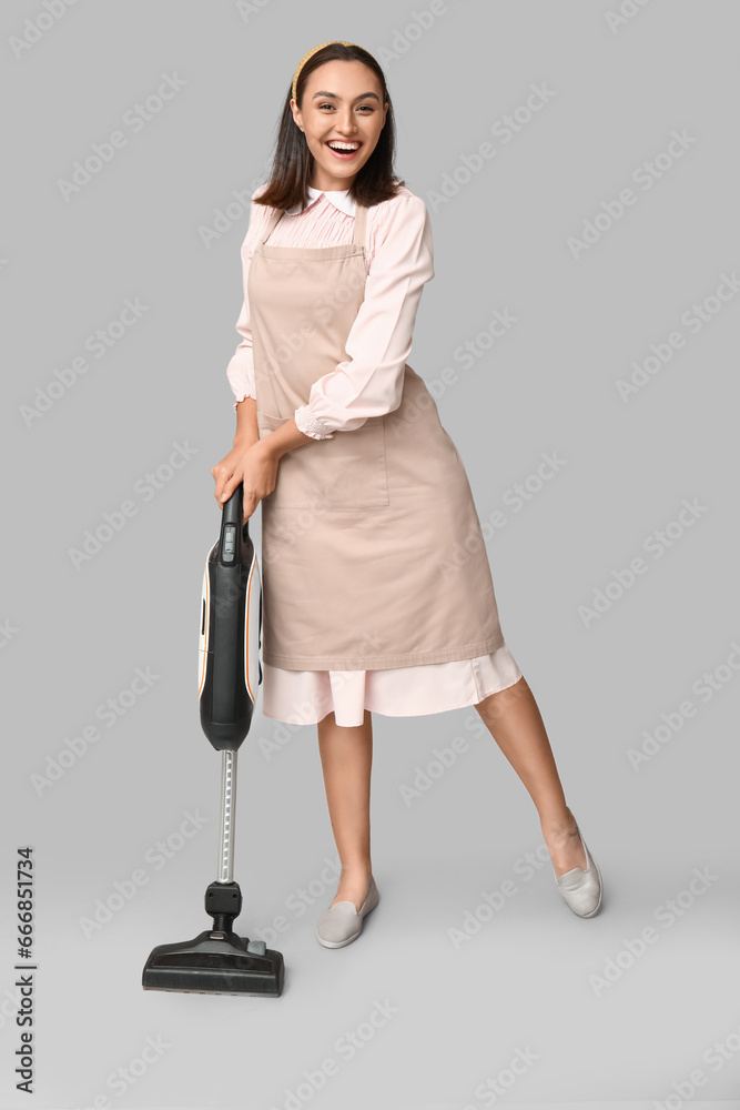 Portrait of happy young housewife in apron with vacuum cleaner on grey background