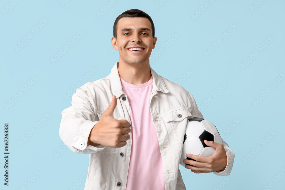 Handsome young man with soccer ball showing thumb-up gesture on blue background