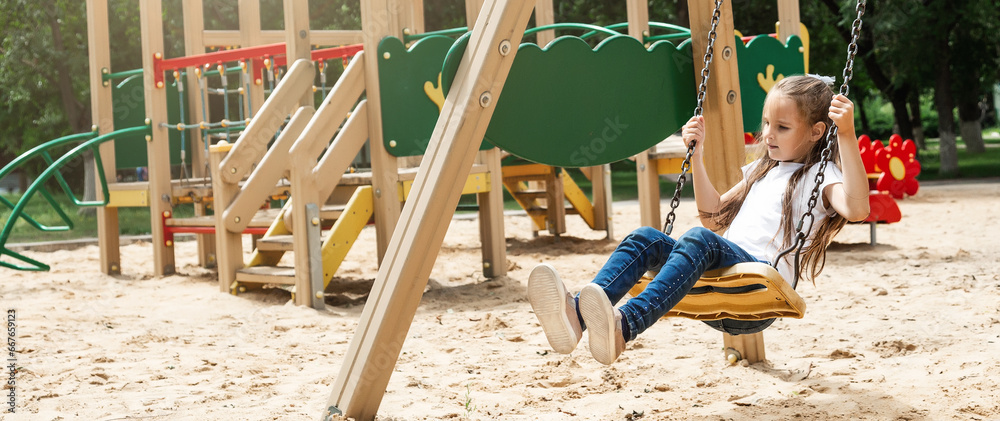 The child plays in the outdoor playground. Active little girl swings on a colorful swing. Healthy summer activity for kids.