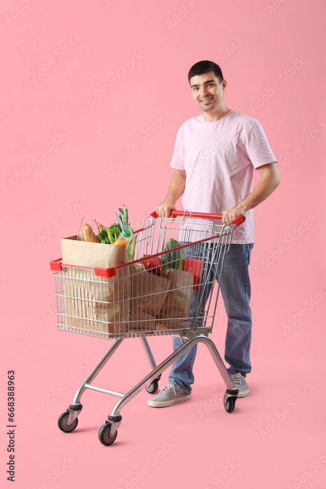Young man with shopping cart and grocery bags on pink background