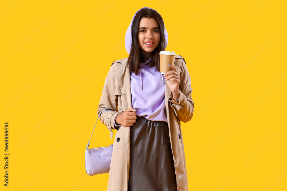 Beautiful young woman in stylish hoodie with cup of coffee on yellow background