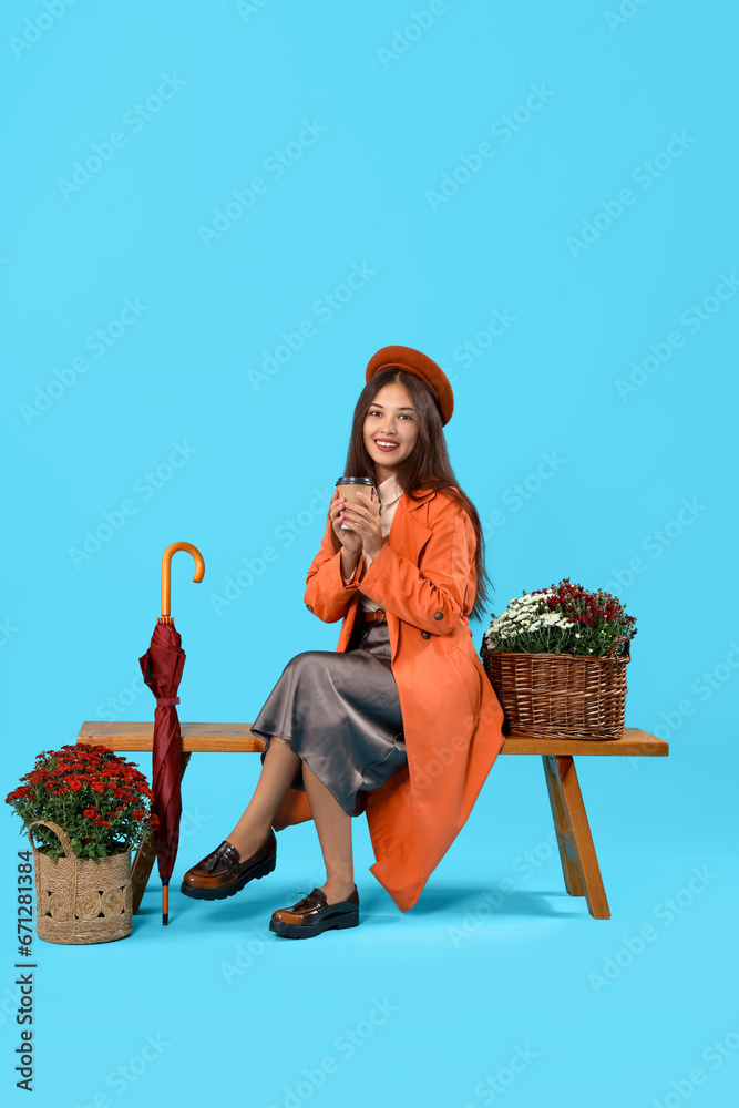 Young Asian woman with coffee cup, chrysanthemum flowers and umbrella sitting on blue background