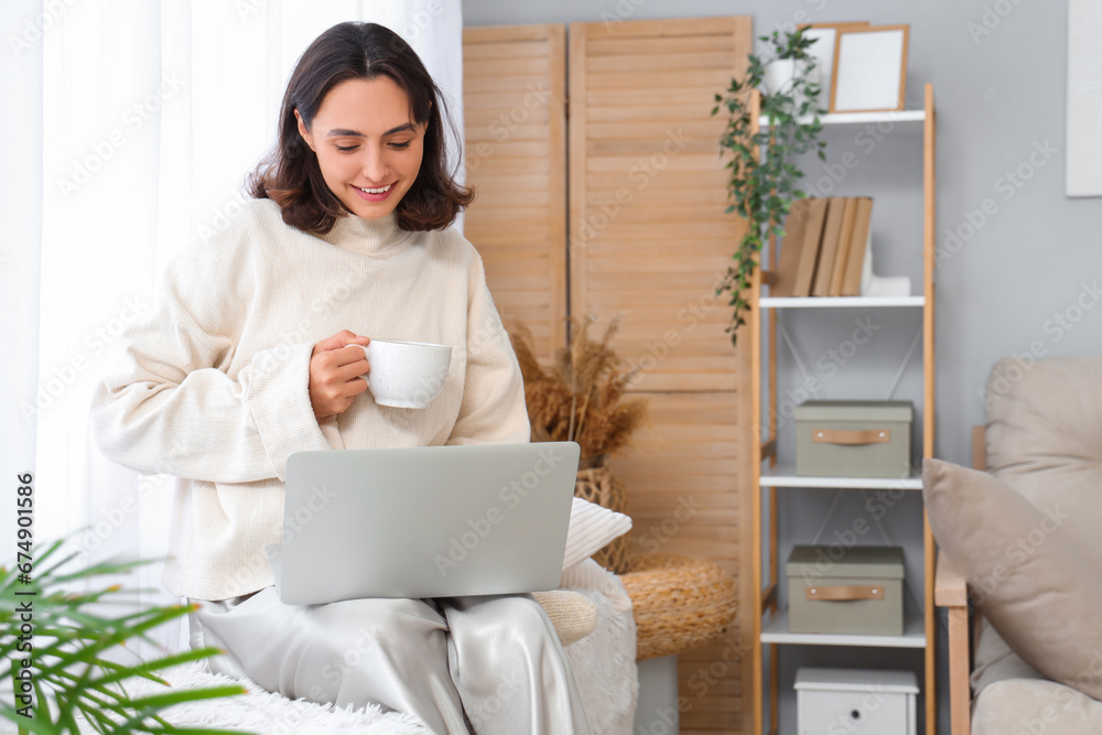 Young woman in warm sweater with cup of tea and laptop at home