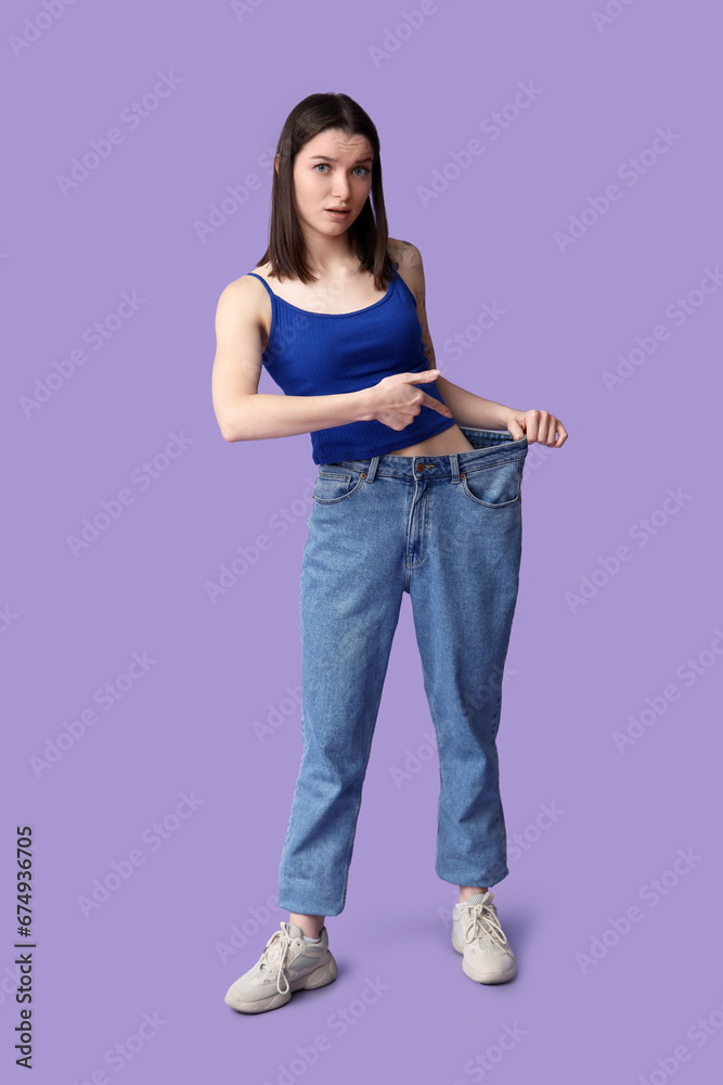 Beautiful young sporty woman pointing at loose jeans on purple background. Weight loss concept