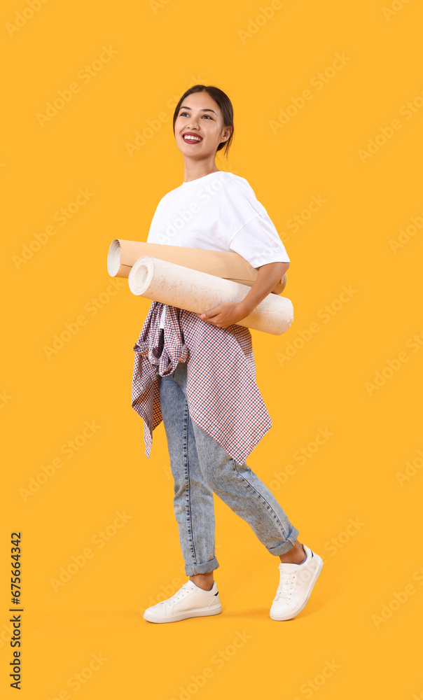 Young Asian woman with wallpaper rolls on yellow background