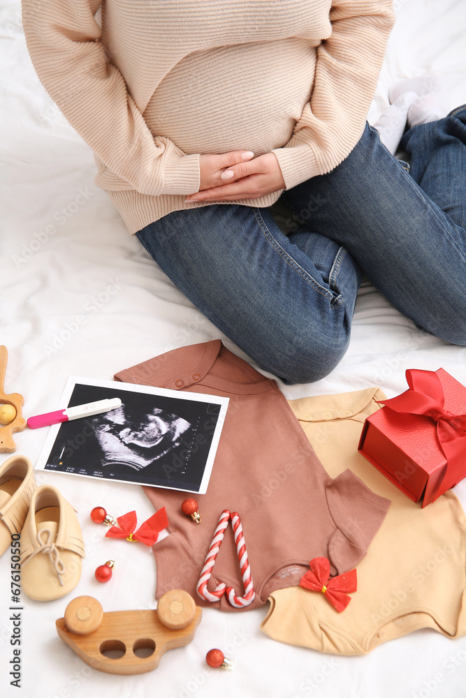 Pregnant woman with baby clothes, sonogram image and Christmas decorations on bed