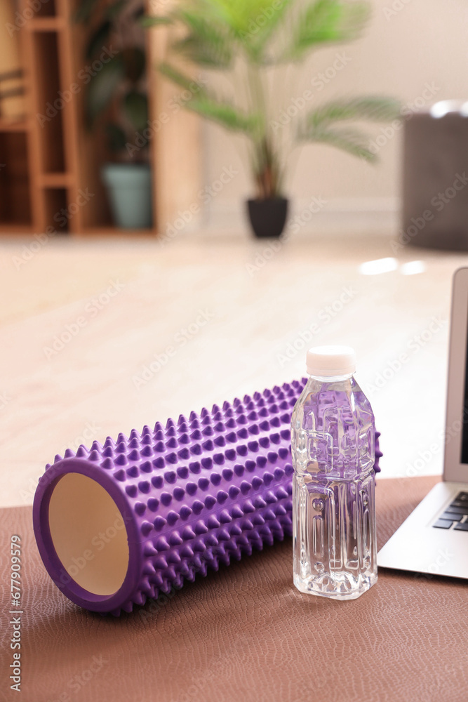 Bottle of water with foam roller on fitness mat in room, closeup