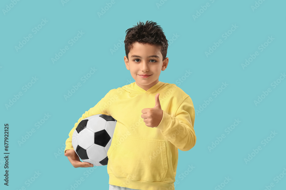 Cute little boy with soccer ball showing thumb-up on blue background