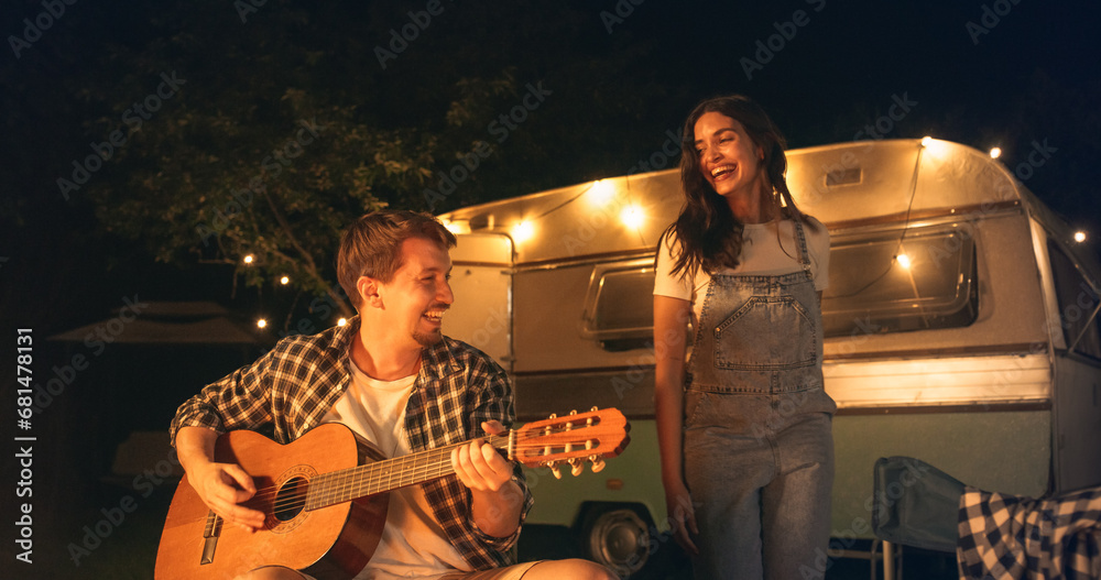 Couple in a Relationship Enjoying a Summer Evening at a Caravan Camping Area. Handsome Boyfriend Playing Acoustic Guitar Next to a Campfire, His Girlfriend is Dancing Barefoot Full of Joy
