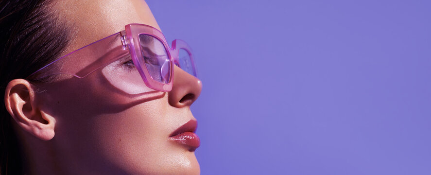 Profile portrait of a beautiful model in trendy square pink sunglasses with glossy make up posing against violet background.  Stylish glasses. Glance glamour picture. Summer Eye wear fashion ..