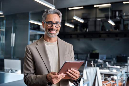 Portrait of happy smiling 45 years old business man executive standing in office using digital tablet. Middle aged businessman manager wearing suit glasses working on professional financial project.