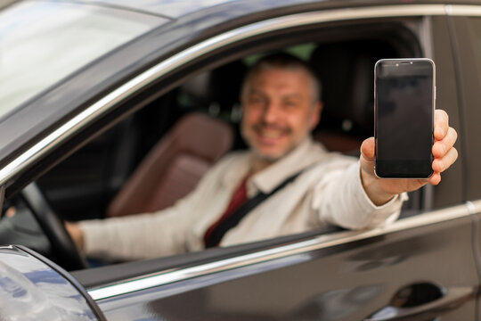 Navigator app. Happy man sitting in car and showing smartphone with empty screen, advertising navigation mobile app, selective focus on phone