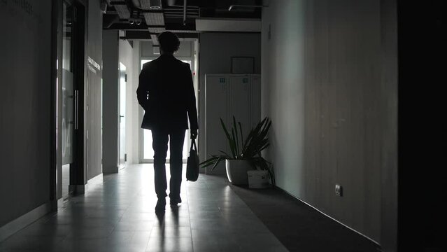 Rear view. The silhouette of a businessman in formal suit walks along a dark corridor of an office building. Confident entrepreneur or manager with a briefcase goes to the exit of the business center