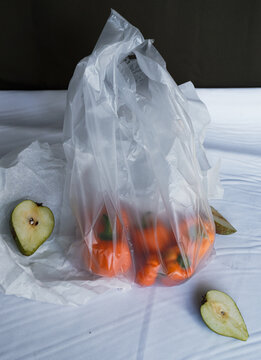 一个透明塑料袋里装着橙色辣椒。包装袋部分打开，放在画面中央。白色布上放着绿色的半个梨子，围绕在袋子周围
