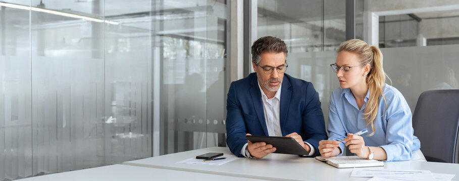 两名专业商务人士协同工作。成熟男女伙伴在办公室讨论项目。团队领导使用平板电脑向实习生讲解工作流程。横幅，留白；