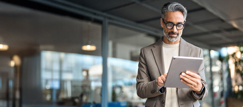 忙碌的成熟资深商业人士、高管站在办公室使用数字平板电脑。中年职业商人、企业经理身着西装，戴着眼镜，手持平板电脑处理金融项目