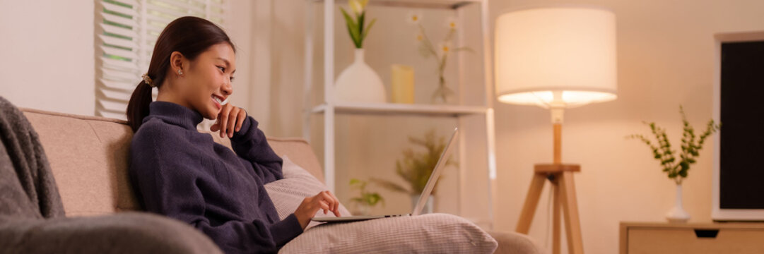 亚洲成年女性微笑着在舒适的家居客厅环境中远程开展线上业务，通过笔记本电脑高效完成工作任务，展现现代生活方式与数字连接带来的居家舒适感