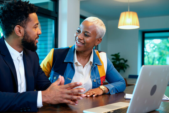 一位年轻的黑人男性和一位年轻的黑人女性坐在桌前讨论商业战略，面带微笑并进行眼神交流，在现代办公室环境中可以看到打开的笔记本电脑和文件。