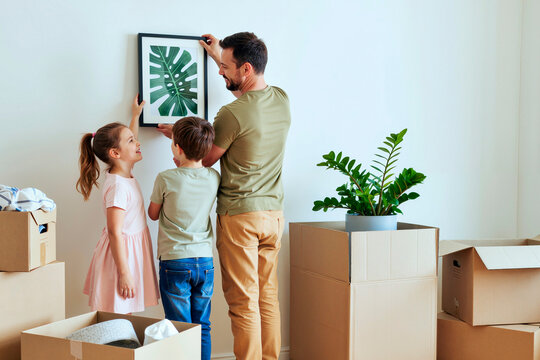 一位高加索中年男子微笑着将一幅装裱好的植物画挂在墙上，一个高加索女孩和一个高加索男孩在一旁看着，周围有搬家纸箱和盆栽植物