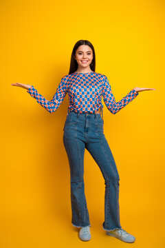 Bright young woman with patterned top and jeans stands against a yellow background smiling with arms open friendly confident lifestyle shot
