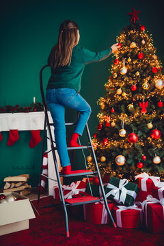 A cheerful helper climbs a ladder to decorate a tall Christmas tree in a cozy living room full of lights gifts and warm holiday atmosphere perfect for a family celebration