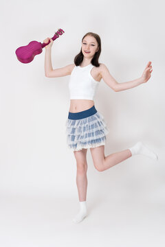 Positive playful young woman ukulelist and cheerleader holding ukulele in her hand and dancing on move. She's pampering, smiling and looking at camera. Full length shot on white background