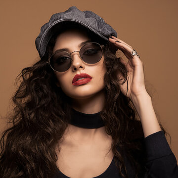Fashion style makeup young woman posing on black sunglasses and summer grey cap, bright red lipstick in studio on brown background
