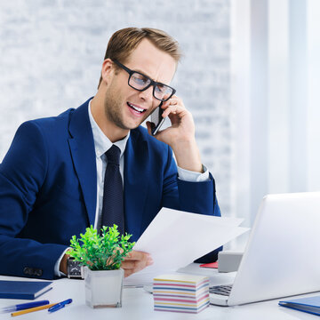 戴着眼镜、身着蓝色夹克的快乐微笑自信商人在办公室使用手机与笔记本电脑工作的肖像照，商业工作概念图，方形构图