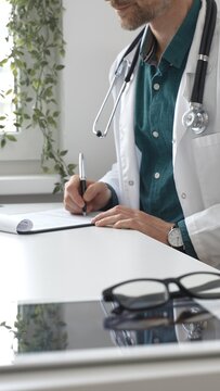 一位穿着绿色衬衫搭配实验室工作服、戴着听诊器的男医生，正坐在办公桌前写医疗处方或填写医疗文件。医药与医疗保健概念