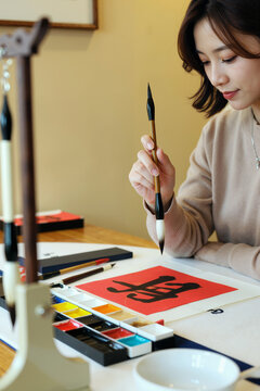 Asian young adult woman practicing calligraphy, holding brush and painting Chinese character on red paper, sitting at table with watercolor paints and art supplies visible in foreground