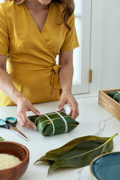 一位高加索年轻成年女性在厨房台面上用绳子将食物包裹在香蕉叶中，双手小心地折叠着叶子，正在准备一道传统菜肴，附近能看到食材。