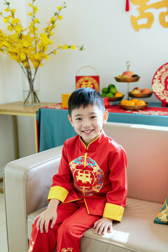 亚洲小男孩身着传统服饰坐在沙发上对着镜头微笑的肖像，背景中有装饰的桌子和水果摆盘，正在庆祝节日