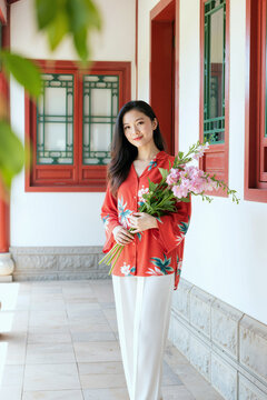 户外站在有着红色窗框和红色装饰的历史建筑旁手持粉色花束对着镜头微笑的年轻亚洲女性肖像