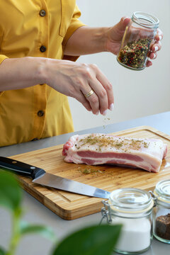 一位高加索女性在木制案板上用混合香料给生五花肉调味，手持玻璃罐撒上香草，为烹饪准备肉类，厨房场景清晰可见