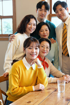 室内六名亚洲中青年摆姿势合影，面带微笑，三名女性坐在放着一杯水的木桌旁，三名男性站在她们身后