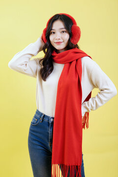 年轻亚洲女性对着镜头微笑的肖像，戴着耳罩和围巾，站在黄色背景前，手摸头部，神情愉悦