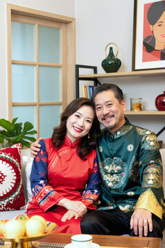 中年亚洲夫妇紧挨着坐在一起对着镜头微笑，女士双手放在腿上，男士手臂环绕着她的肩膀，能看到传统服饰，家庭室内背景