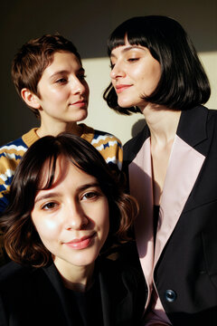 三名年轻的白人女性紧挨着摆姿势的肖像照，其中两人相互对视，一人看向镜头，在明亮的室内环境中展现出自然的表情