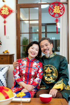 中年亚洲夫妇身着传统服饰，坐在客厅沙发上，面带微笑对着镜头，在布置有传统装饰的客厅里庆祝节日，装饰上写着“幸福”字样。