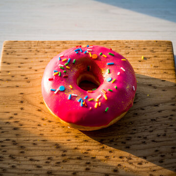 清晨阳光下浅木色案板上带糖珠的亮粉色糖霜甜甜圈