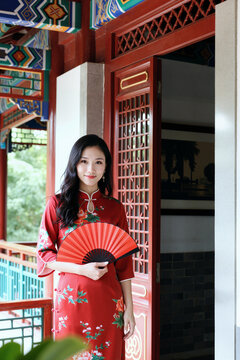 年轻亚洲成年女性站在传统阳台上手持折扇的肖像，面带微笑看向镜头，身着刺绣服饰，可见复杂的建筑细节