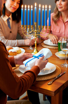 一群来自不同种族的年轻男女正在庆祝光明节，他们围坐在摆放着烛台的餐桌旁，手持用系着蓝色丝带的餐巾包裹的礼物，一同享用节日大餐。