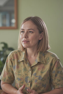 年轻的白人成年女性坐在桌前的肖像，微微看向镜头外，表情平静，双手在前景可见，室内场景，背景柔和。