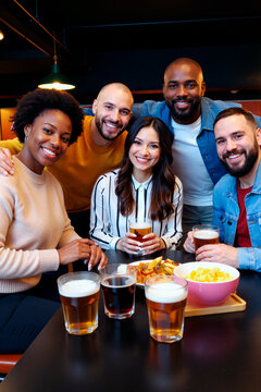 一群年轻的多民族朋友微笑着围坐在桌旁，手持饮品和小吃，看着镜头，在休闲社交场合露出愉悦的表情