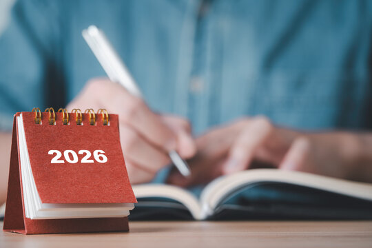 展示一本 2026 年桌面日历，上面有一个人正在笔记本上书写，体现了在商业和个人生活中为新的一年进行规划、组织、设定未来目标、时间管理和做准备的场景