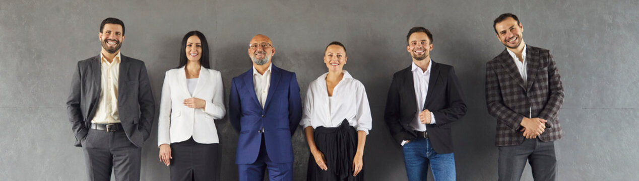 快乐自信的专业商务人士团队在工作室墙边成排站立的团体肖像。穿着正式西装的企业员工、企业家共同摆姿势。人力资源概念；