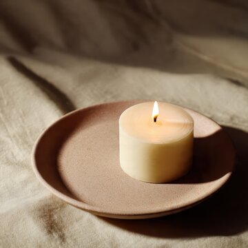 A lit candle on a pink plate sits atop a wrinkled white surface casting a soft shadow in the dim light